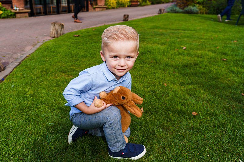 A young boy with blonde hair, wearing a light blue shirt and gray pants, crouches on a grassy lawn while holding a brown stuffed animal. The background shows a pathway, some wooden structures, and another person walking away. Image by Documentary Wedding Photographer Lewis Bishop Photography.