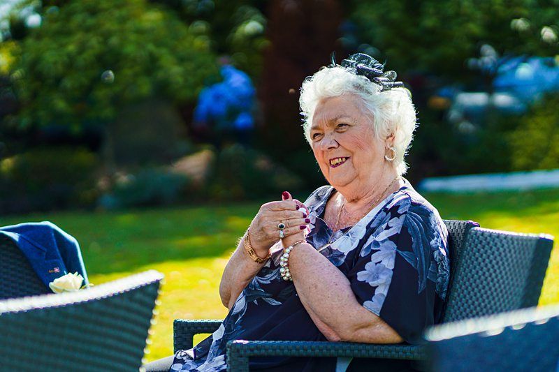 An elderly woman with white hair sits outdoors on a wicker chair. She is smiling and wearing a floral-patterned dress, beaded bracelets, and a fascinator. The sun is shining, and there are green trees in the background. Image by Documentary Wedding Photographer Lewis Bishop Photography.