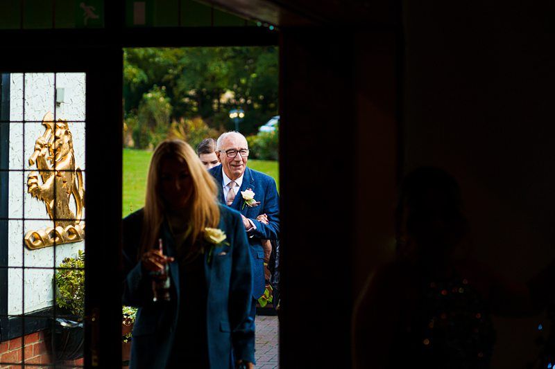 A group of people enter a dimly lit room through a door with stained glass windows. In focus is an older man in a suit and glasses, smiling. In the foreground, a woman holds a drink and wears a flower on her jacket. Outside, greenery is visible. Image by Documentary Wedding Photographer Lewis Bishop Photography.