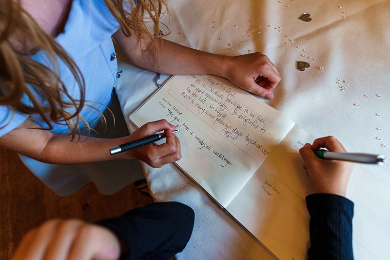 Two people are seen from above writing in a guestbook on a table adorned with small decorative hearts. One person in a blue outfit holds a pen poised to write, while another person wearing black is writing. The guestbook is open, displaying handwritten messages. Image by Documentary Wedding Photographer Lewis Bishop Photography.