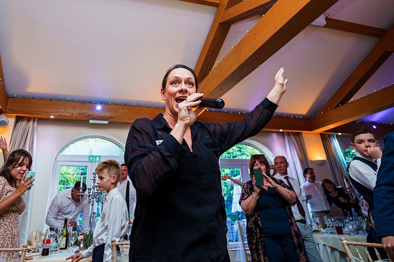 A person holds a microphone and raises one hand while speaking or singing in a lively room filled with people. Some individuals are seated at tables, while others stand, listening and watching attentively. Strings of lights decorate the wooden beams above. Image by Documentary Wedding Photographer Lewis Bishop Photography.