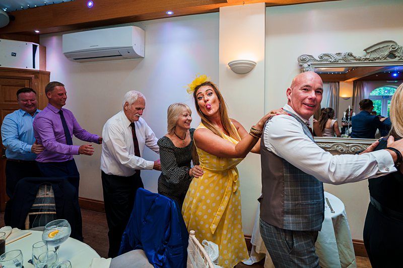A group of people in formal attire form a conga line inside a decorated room. A woman in a yellow dress and matching fascinator is at the front, playfully making a face. The other participants are smiling and enjoying the moment. A mirror and air conditioner are on the walls. Image by Documentary Wedding Photographer Lewis Bishop Photography.