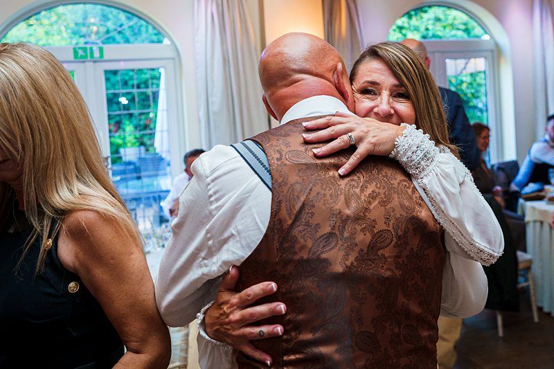 A couple is embracing warmly at an indoor gathering. The man, wearing a brown patterned vest and white shirt, has his back to the camera, while the woman, dressed in white, smiles with her eyes closed. Other guests are visible in the background, engaged in conversation. Image by Documentary Wedding Photographer Lewis Bishop Photography.