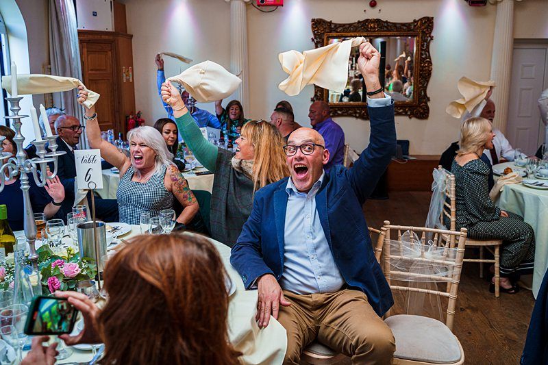 Guests at a celebratory event joyously wave napkins in the air while seated at banquet tables. A man in the foreground, wearing glasses and a blue blazer, expresses excitement, while other smiling attendees join in the lively festivities. Image by Documentary Wedding Photographer Lewis Bishop Photography.