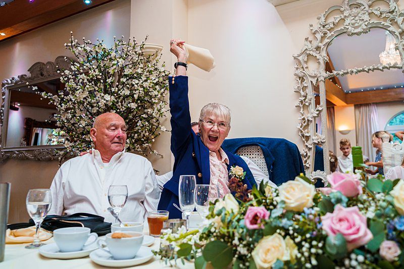 An elderly woman, smiling and jubilant, raises her hand in celebration at a decorated banquet table. An elderly man sits calmly beside her. The table is adorned with flowers, teacups, and glassware. In the background, ornate mirrors and floral arrangements can be seen. Image by Documentary Wedding Photographer Lewis Bishop Photography.