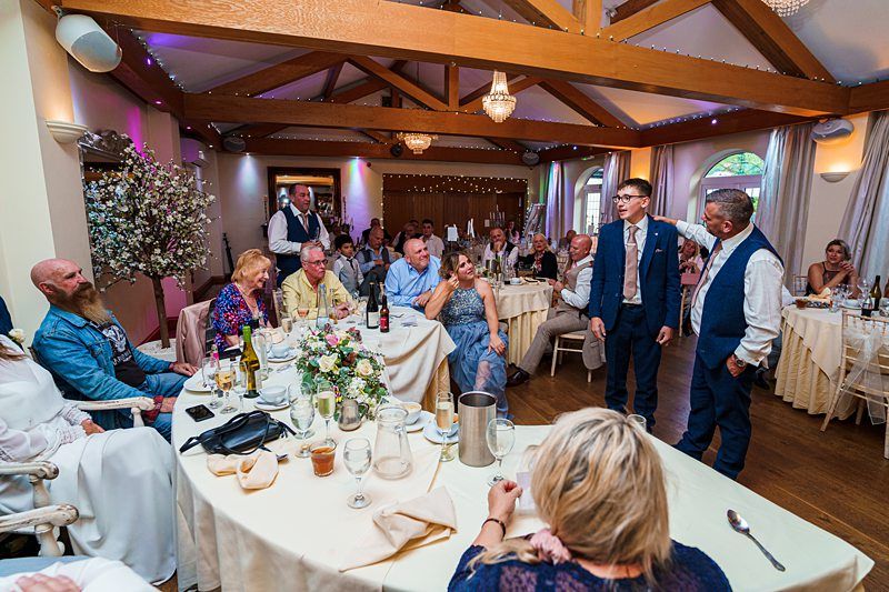 A lively wedding reception in a warmly lit hall. Guests are seated at round tables adorned with floral centerpieces and drinks. Two men stand in the center, one gesturing animatedly while others watch and smile. Wooden beams and draped lights add charm to the atmosphere. Image by Documentary Wedding Photographer Lewis Bishop Photography.