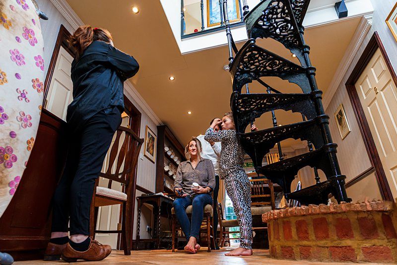 Three people are indoors near a black spiral staircase. A woman is sitting on a chair, smiling, while a person in black operates a camera. A child in patterned clothing is holding onto the staircase railing. The setting appears casual and home-like. Image by Documentary Wedding Photographer Lewis Bishop Photography.