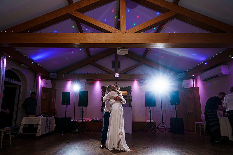 A bride and groom share a romantic dance under wooden beams adorned with string lights. Colorful lighting spots illuminate the scene, while a few guests look on from a dimly lit background. Two bright lights and speakers are set up nearby, enhancing the celebratory atmosphere. Image by Documentary Wedding Photographer Lewis Bishop Photography.