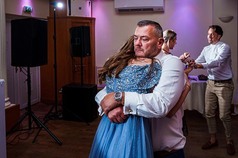 A man in a white shirt hugs a young girl in a sparkly blue dress on a dance floor with dim lighting and a purple glow. Other people can be seen in the background, some adjusting equipment, giving a feeling of a celebratory event or party. Image by Documentary Wedding Photographer Lewis Bishop Photography.