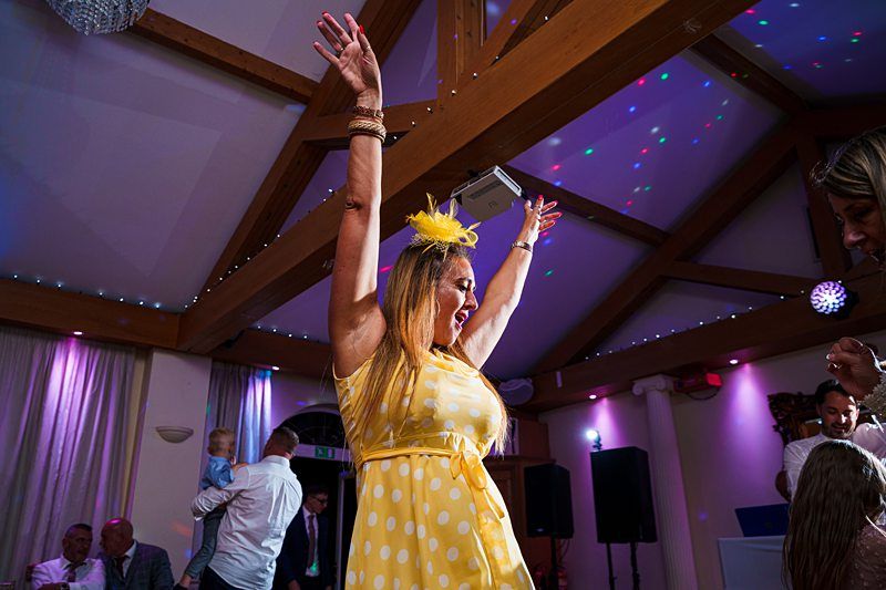 A woman in a yellow polka dot dress and matching yellow fascinator dances with her arms raised in a room decorated with colorful lights. Other people can be seen casually enjoying the event in the background. Image by Documentary Wedding Photographer Lewis Bishop Photography.