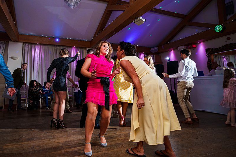 People are dancing joyfully in a room with wooden beams and colorful lighting. A woman in a vibrant pink dress and another in a yellow dress are at the center. Other guests, dressed in a variety of outfits, are also dancing or sitting in the background, enjoying the event. Image by Documentary Wedding Photographer Lewis Bishop Photography.