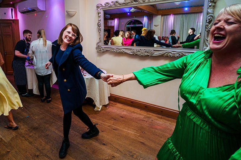 Two women are joyfully dancing hand-in-hand in a warmly lit room with a wooden floor. One wears a navy coat and the other a bright green dress. Guests and a large mirror reflecting the scene are visible in the background. The atmosphere appears festive and lively. Image by Documentary Wedding Photographer Lewis Bishop Photography.