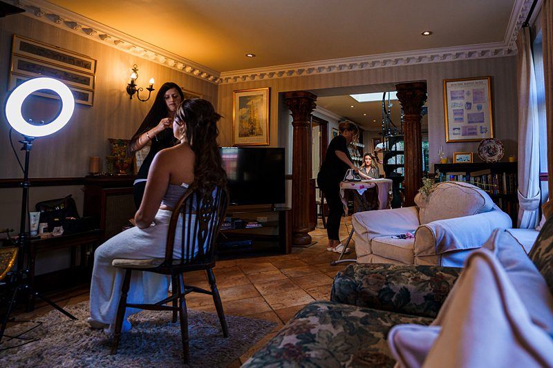 A bride is getting her makeup done by a makeup artist in a cozy, furnished room with soft lighting. Another person is getting ready in the background. The space is decorated with framed pictures, a plush sofa, a standing mirror, and wooden columns. Image by Documentary Wedding Photographer Lewis Bishop Photography.