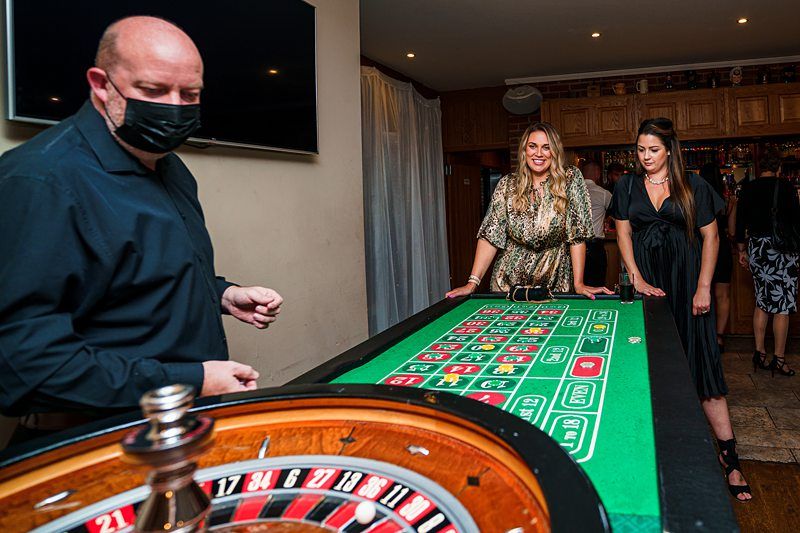 A man wearing a mask is operating a roulette table, while two women stand at the table watching and waiting to place their bets. One woman is wearing a patterned dress, and the other is in a black dress. The setting appears to be indoors, with subdued lighting. Image by Documentary Wedding Photographer Lewis Bishop Photography.