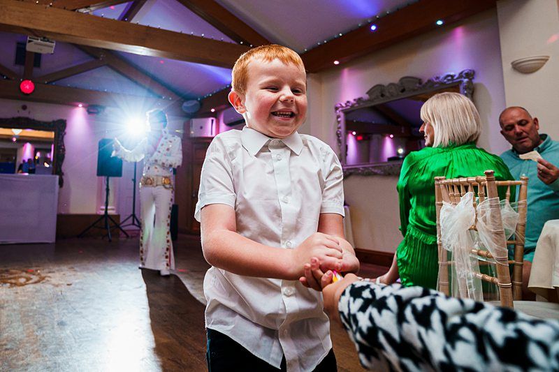 A young boy with short red hair and wearing a white shirt smiles joyfully while dancing at a lively event. In the background, adults are also enjoying themselves, with one in a green outfit and another in a white suit. Colorful lights add to the festive atmosphere. Image by Documentary Wedding Photographer Lewis Bishop Photography.