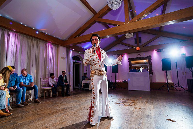 Person dressed in a white Elvis Presley-style jumpsuit, complete with a cape, sunglasses, and a microphone, performing in a room with a wooden floor and vaulted ceiling. A few seated people are in the background, and colored lights illuminate the scene. Image by Documentary Wedding Photographer Lewis Bishop Photography.