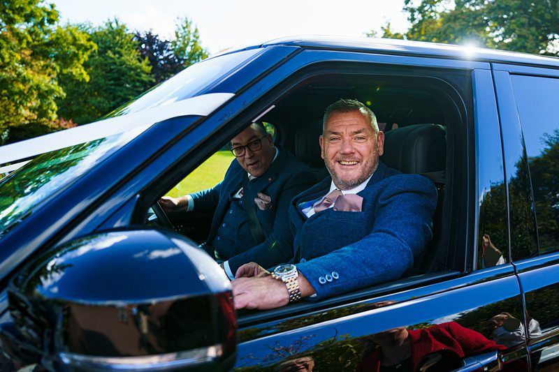 Two smiling men in formal attire, one driving and the other in the passenger seat of a sleek black vehicle. The driver wears a bow tie and watch. They appear ready for a special occasion, with sunlight filtering through lush greenery in the background. Image by Documentary Wedding Photographer Lewis Bishop Photography.