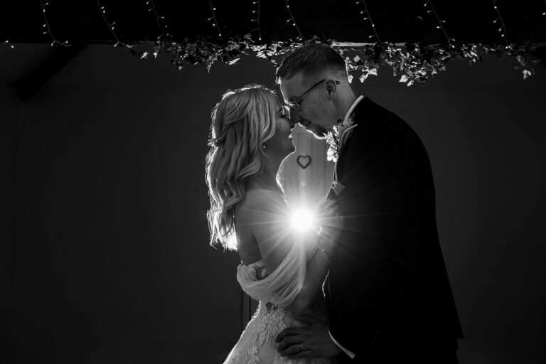 A bride and groom stand face to face in a dimly lit setting, with the sun shining between them, creating a heart-shaped reflection on the lens. Image by Documentary Wedding Photographer Lewis Bishop Photography.