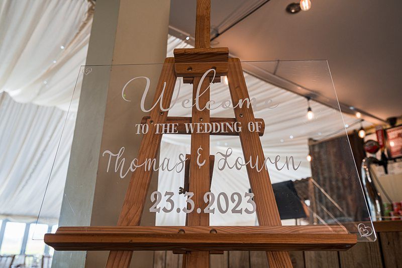 A clear acrylic wedding sign on a wooden easel reads "Welcome to the wedding of Thomas & Lowan, 23.3.2023," set in a decorated indoor venue. Image by Documentary Wedding Photographer Lewis Bishop Photography.