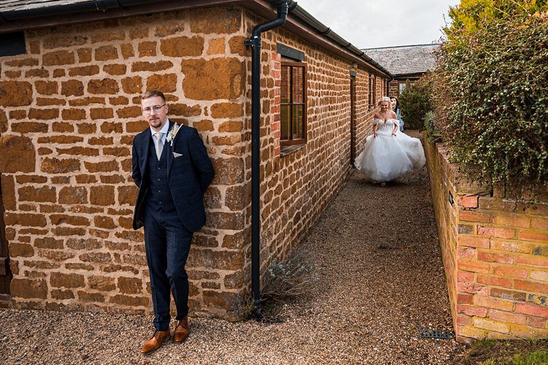A groom in a dark suit stands by a brick building while a bride in a white gown walks towards him on a path. Image by Documentary Wedding Photographer Lewis Bishop Photography.