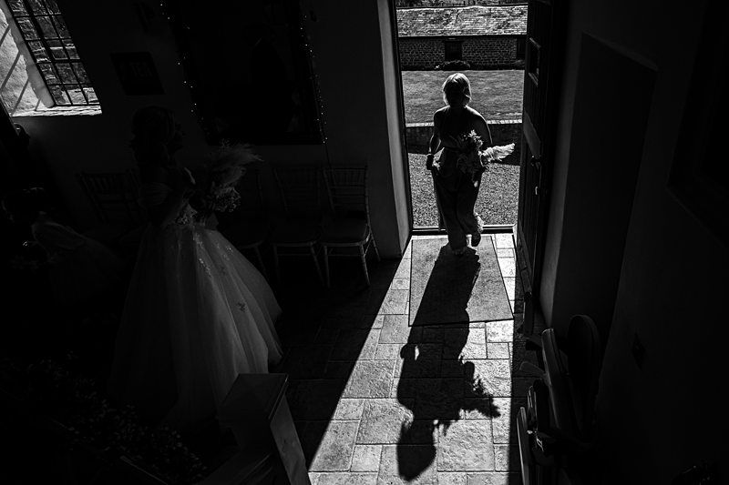 A person holding flowers walks through a sunlit doorway casting a long shadow on the floor while another person in a wedding dress stands inside a dimly lit room. Image by Documentary Wedding Photographer Lewis Bishop Photography.
