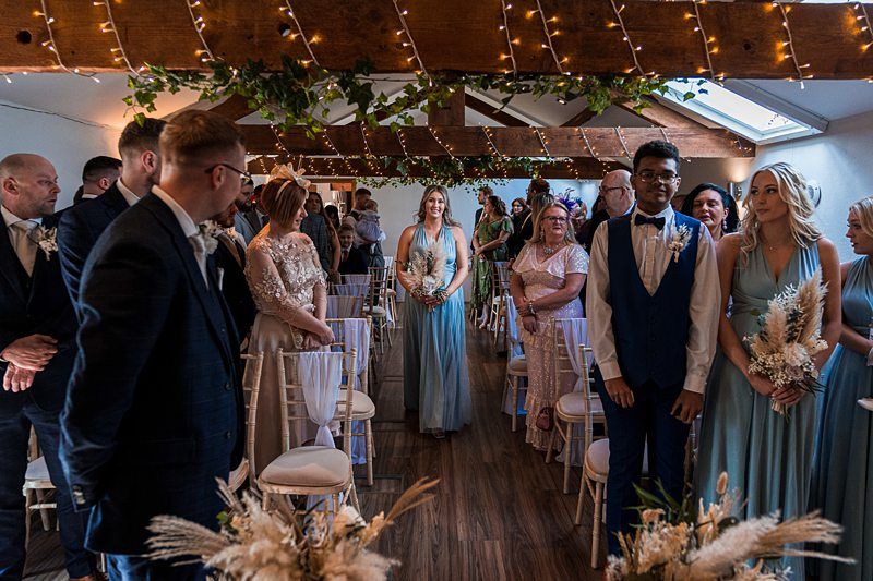 A bride walks down the aisle during a wedding ceremony in a decorated venue, while guests stand and observe. The ceiling is adorned with greenery and string lights. Image by Documentary Wedding Photographer Lewis Bishop Photography.