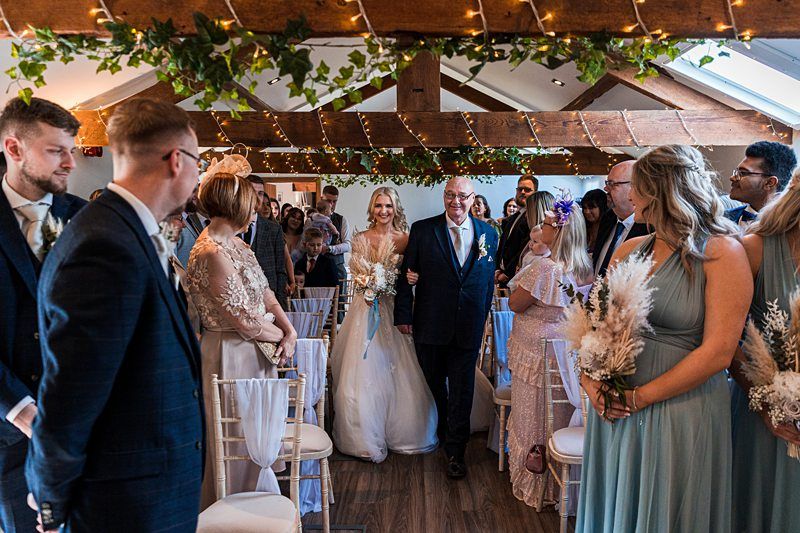 A bride in a white gown walks down an aisle decorated with greenery and lights, accompanied by an older man. Guests on either side of the aisle turn to watch. Image by Documentary Wedding Photographer Lewis Bishop Photography.