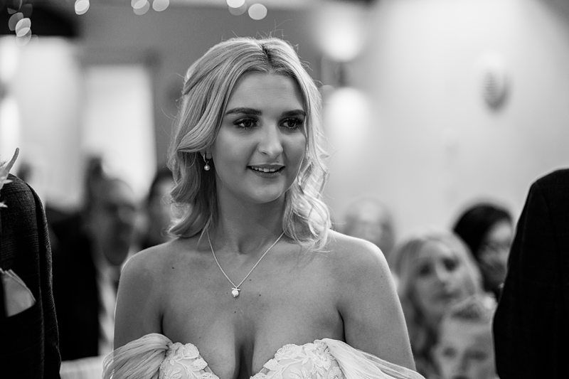 A woman in a wedding gown stands indoors, facing forward, with people seated in the background. The image is in black and white. Image by Documentary Wedding Photographer Lewis Bishop Photography.
