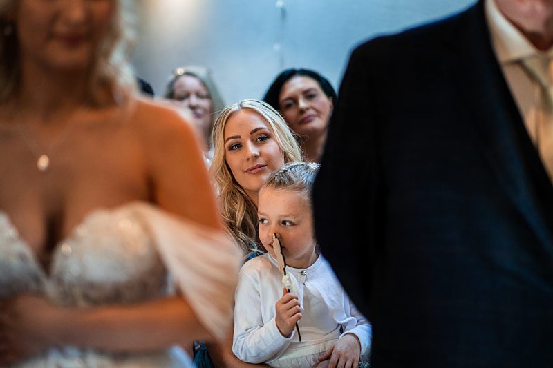 A woman smiles while holding a small child who is looking ahead, surrounded by other people in the background during what appears to be a formal event. The focus is on the background figures. Image by Documentary Wedding Photographer Lewis Bishop Photography.