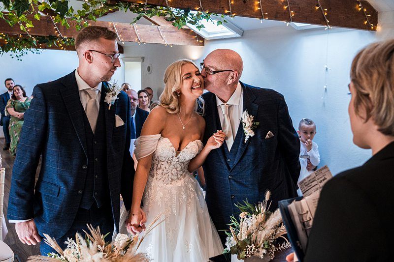 A bride smiles while holding hands with a groom as an older man kisses her cheek. People are watching in the background. The venue is decorated with greenery and lights. Image by Documentary Wedding Photographer Lewis Bishop Photography.