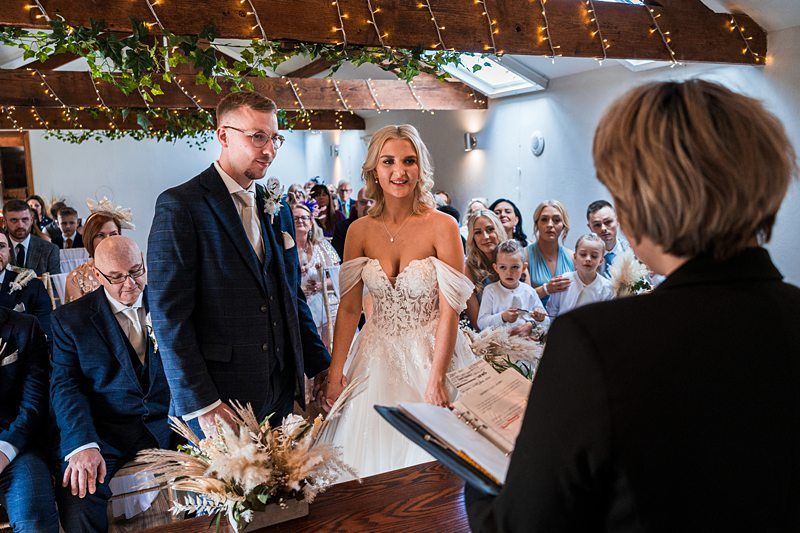 A bride and groom stand at the altar during their wedding ceremony. The officiant is reading from a book while guests watch from their seats. The venue is decorated with greenery and fairy lights. Image by Documentary Wedding Photographer Lewis Bishop Photography.