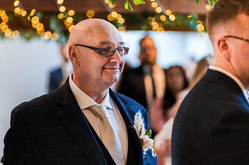 Elderly man in a suit and tie smiles at a gathering, adorned with string lights and greenery. Image by Documentary Wedding Photographer Lewis Bishop Photography.