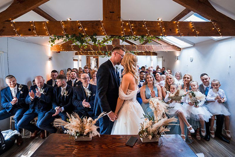A couple faces each other holding hands in a well-lit room filled with guests seated on both sides. The room is decorated with string lights and greenery. People are clapping and smiling. Image by Documentary Wedding Photographer Lewis Bishop Photography.