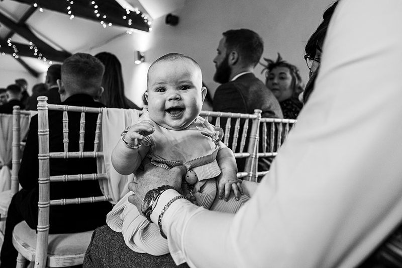 A baby smiles while sitting on an adult's lap during an indoor event with people sitting on chairs around. The setting includes overhead string lights and visible chairs. Image by Documentary Wedding Photographer Lewis Bishop Photography.