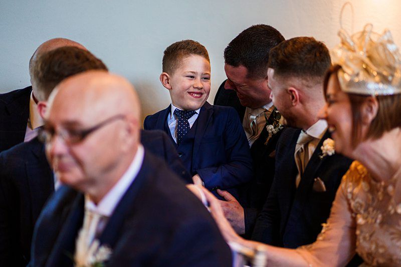 A young boy in a blue suit smiles at two men while a woman in a hat sits beside them at an event. Four other people, including a bald man in glasses, are seated nearby. Image by Documentary Wedding Photographer Lewis Bishop Photography.
