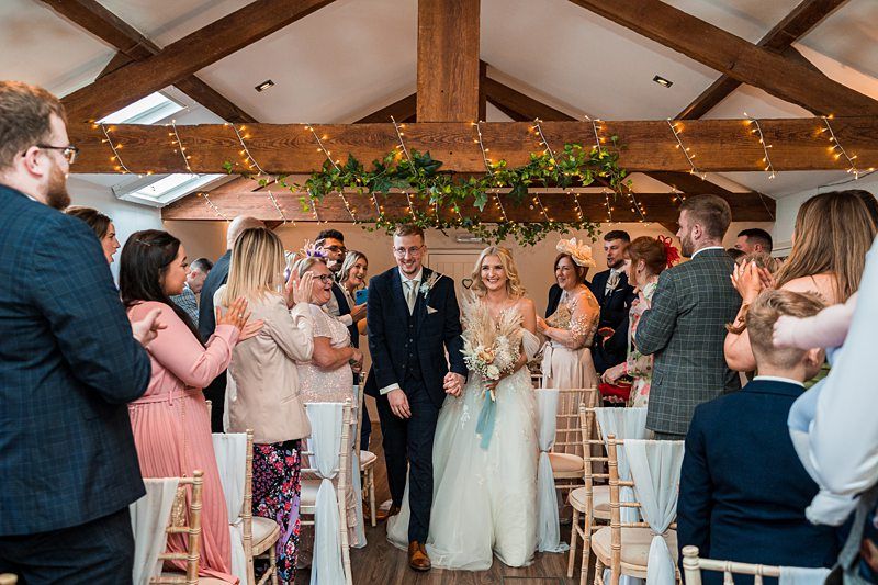 A bride and groom walk down the aisle together in a small, rustic venue decorated with string lights and greenery, surrounded by smiling and clapping guests. Image by Documentary Wedding Photographer Lewis Bishop Photography.