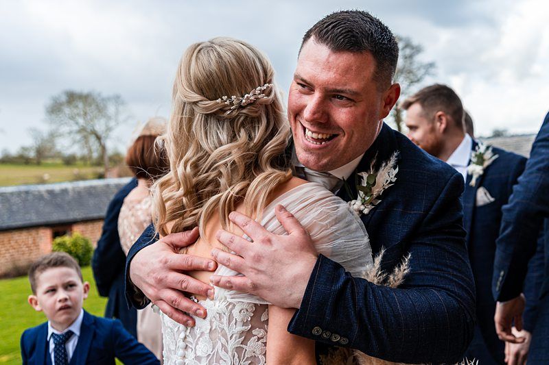 A man in a suit hugs a woman in a white dress, smiling. Another man and a boy in suits stand nearby, with a rural background. Image by Documentary Wedding Photographer Lewis Bishop Photography.