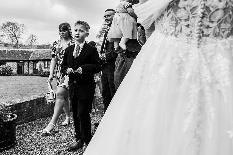 Black and white photo of a young boy in a suit holding something, standing near a woman in a dress, with a wedding dress partially visible on the right. Other individuals and rural scenery in the background. Image by Documentary Wedding Photographer Lewis Bishop Photography.