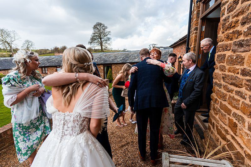 A group of people, dressed in formal attire, are gathered outside a rustic brick building. Some are hugging, and others are conversing. It appears to be a celebratory event, likely a wedding. Image by Documentary Wedding Photographer Lewis Bishop Photography.