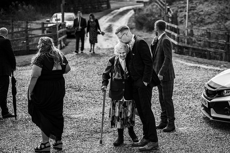 A group of people, dressed formally, are gathered outdoors on a gravel pathway. An elderly woman with a walking stick is at the center, accompanied by a man who is leaning in to talk to her. Image by Documentary Wedding Photographer Lewis Bishop Photography.