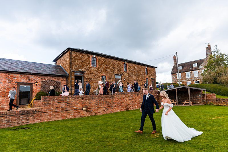 A bride and groom walk through a grassy courtyard with wedding guests gathered near a brick building in the background. Image by Documentary Wedding Photographer Lewis Bishop Photography.