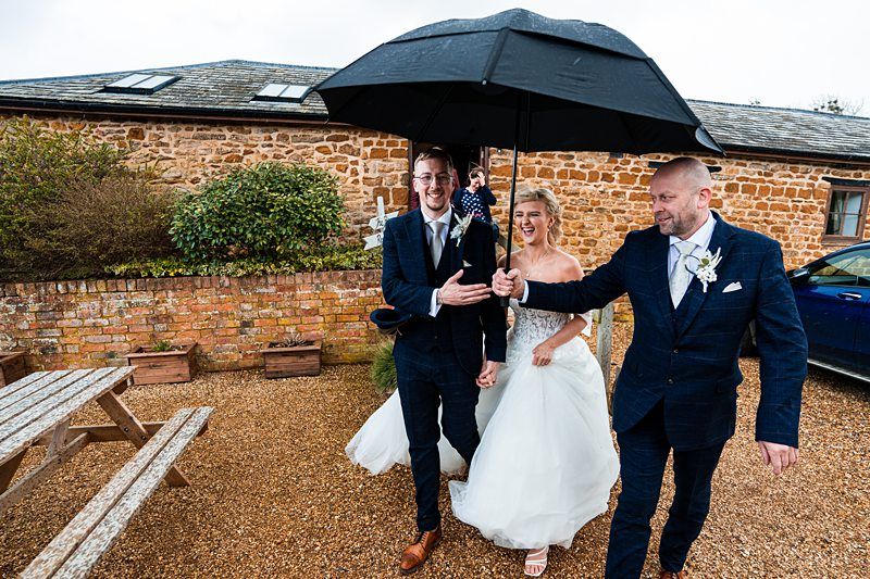 A bride and groom, dressed in formal wedding attire, walk outdoors under a large black umbrella held by another man in a suit on a rainy day. They are smiling and appear joyful. Image by Documentary Wedding Photographer Lewis Bishop Photography.