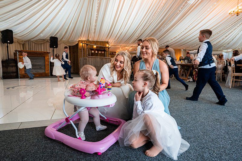 A baby in a pink walker, a child in a white dress, and two women smile while interacting on the floor of a decorated, tent-like venue with other guests in the background. Image by Documentary Wedding Photographer Lewis Bishop Photography.