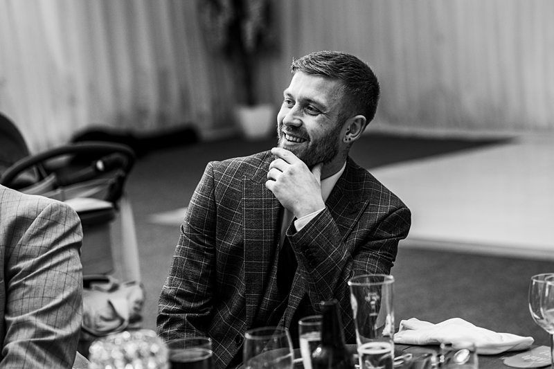 A man in a plaid suit jacket sits at a table, smiling and holding his chin. The background appears to be a room with draped curtains and a high chair is visible. Image by Documentary Wedding Photographer Lewis Bishop Photography.