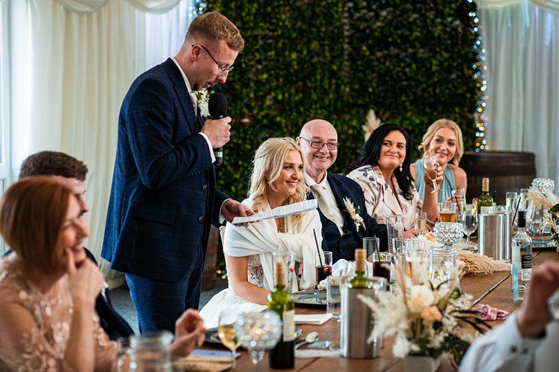 A man in a suit gives a speech using a microphone at a wedding reception. The bride, groom, and other guests are seated at a table, smiling and listening attentively. The background is decorated with greenery. Image by Documentary Wedding Photographer Lewis Bishop Photography.
