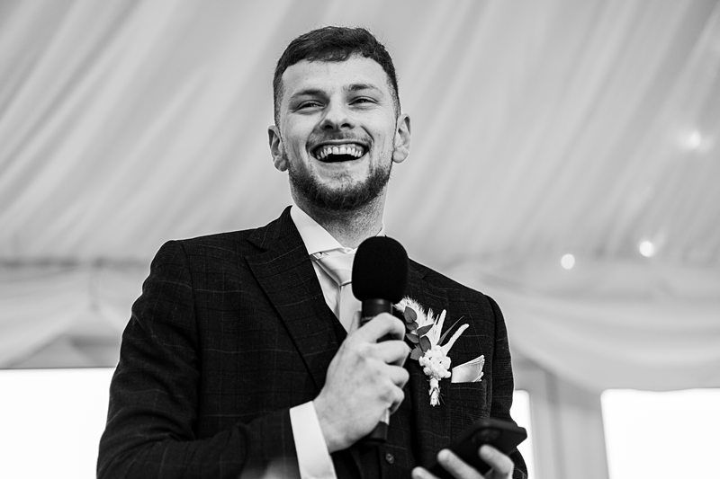 A man in a suit holding a microphone smiles while speaking, with a boutonniere pinned to his jacket. The background features draped fabric. Image by Documentary Wedding Photographer Lewis Bishop Photography.