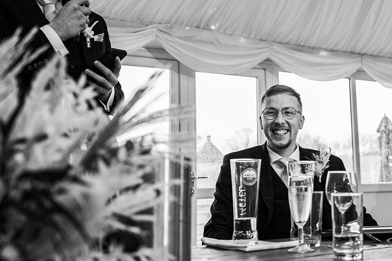 A man in a suit and glasses, seated at a table, smiling broadly during a speech at an indoor event while another person holds a microphone nearby. Image by Documentary Wedding Photographer Lewis Bishop Photography.