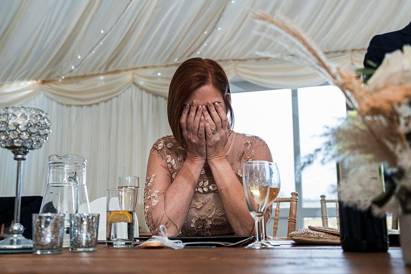 A woman in a beige dress covers her face with her hands while sitting at a table in an elegantly decorated room. Image by Documentary Wedding Photographer Lewis Bishop Photography.