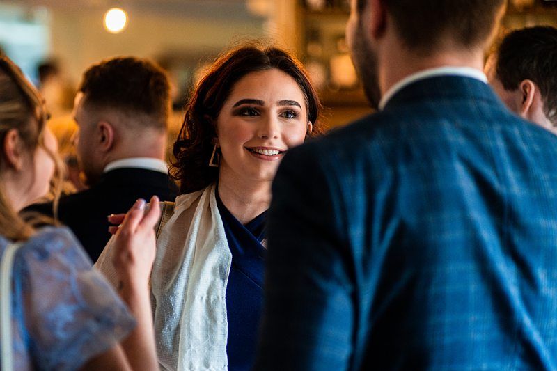 A woman with dark hair and a white scarf is talking to a man in a blue suit at a social gathering. Other people are conversing in the background. Image by Documentary Wedding Photographer Lewis Bishop Photography.