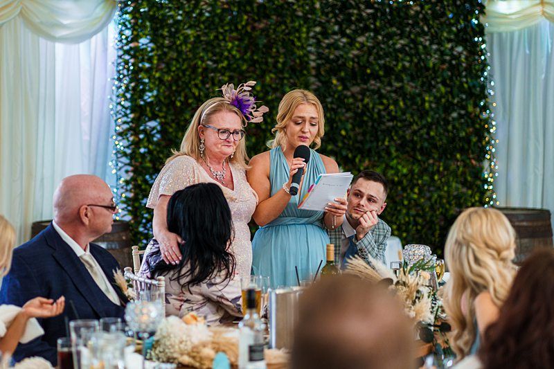 A woman with a microphone speaks while reading from a paper, as another woman stands beside her. People sit around a table in a decorated setting with greenery and lights in the background. Image by Documentary Wedding Photographer Lewis Bishop Photography.